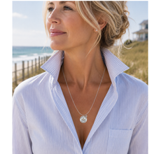 Load image into Gallery viewer, A woman wearing a silvertone round beach tag Beach Haven pendant necklace with a beach-themed engraving, paired with a light blue and white striped button-down shirt, set against a sunny beach background.