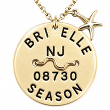 Load image into Gallery viewer, A gold-toned round beach tag pendant necklace engraved with 'BRIELLE NJ 08730 SEASON' and a wavy line, hanging from a chain with a small starfish charm on a white background.