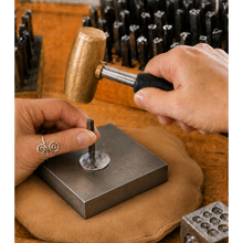 Load image into Gallery viewer, A close-up of a jeweler's hands using a brass hammer to strike a small metal setting on a square steel anvil, with rows of similar hammers and tools arranged on a wooden workbench.