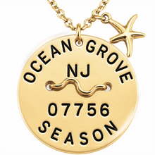Load image into Gallery viewer, A gold-toned round beach tag pendant necklace engraved with 'OCEAN GROVE NJ 07756 SEASON' and a wavy line motif, with a small starfish charm attached at the top, shown on a white background.