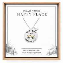 Load image into Gallery viewer, A silver personalized beach badge necklace with a round disc pendant and starfish charm, displayed on a white card inside a kraft box. The card features the text 'WEAR YOUR HAPPY PLACE' and custom text reading 'SEASON NJ 07712 ASBURY PARK'.