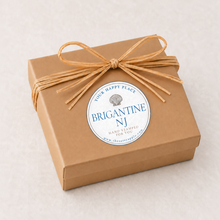 Load image into Gallery viewer, A square gift box wrapped in brown kraft paper and tied with a natural jute ribbon bow, featuring a round white sticker label that reads 'BRIGANTINE NJ' with a seashell graphic, photographed on a light beige textured background.
