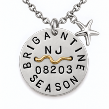 Load image into Gallery viewer, A silver-toned round beach tag pendant necklace with a starfish charm, featuring a gold wave accent and engraved text reading 'BRIGANTINE SEASON' with 'NJ' and a number on a white background.
