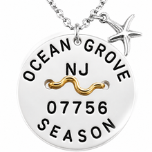 Load image into Gallery viewer, A silver round beach tag pendant necklace engraved with 'OCEAN GROVE NJ 07756 SEASON' and a gold wavy line detail, shown with a silver starfish charm on a white background.

