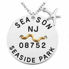 Load image into Gallery viewer, A silver round beach tag pendant necklace engraved with 'SEASON NJ 08752 SEASIDE PARK' and a gold wave detail, featuring a small silver starfish charm on a white background.
